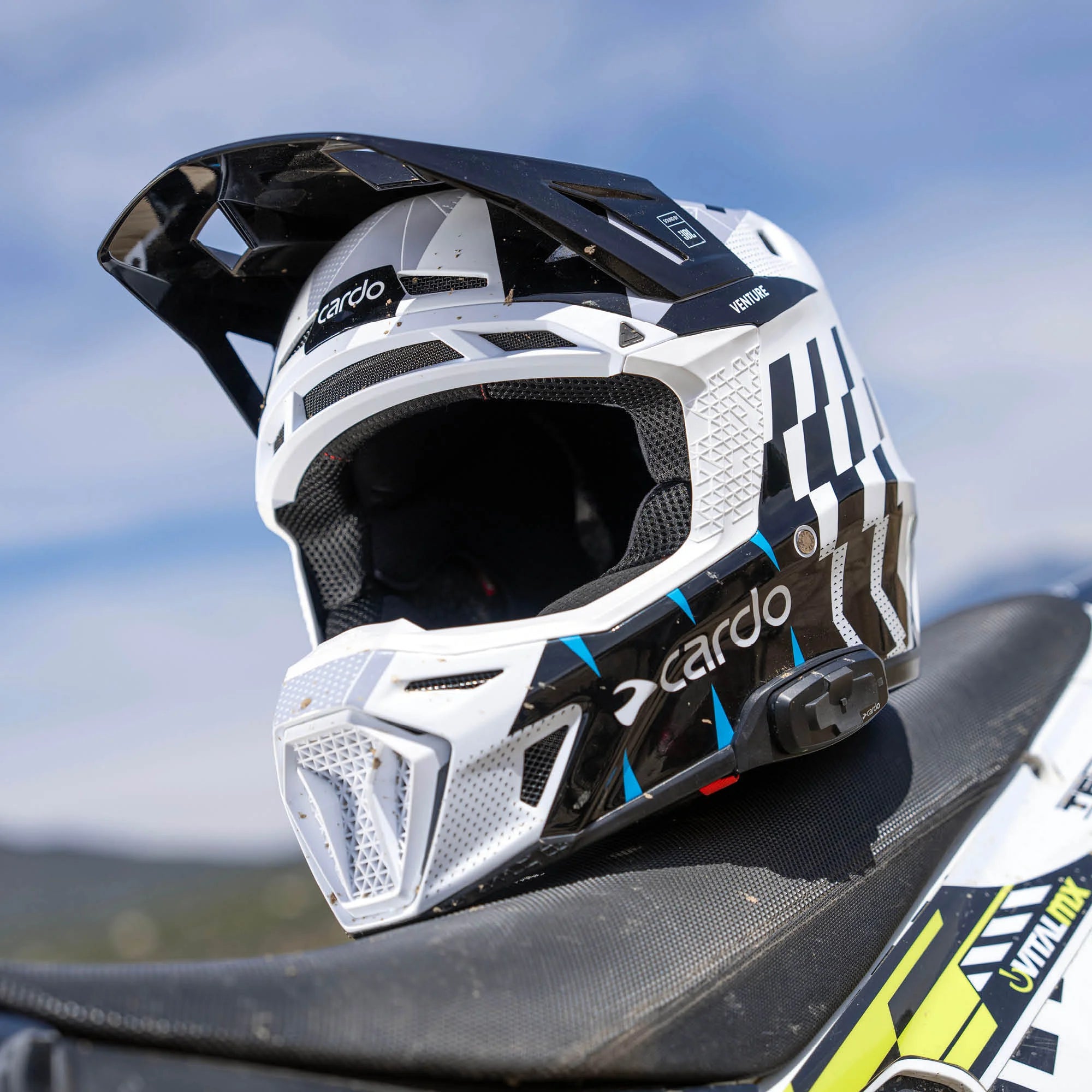 Cardo Venture off-road motorcycle helmet in white and blue, three-quarter view resting on dirt bike seat