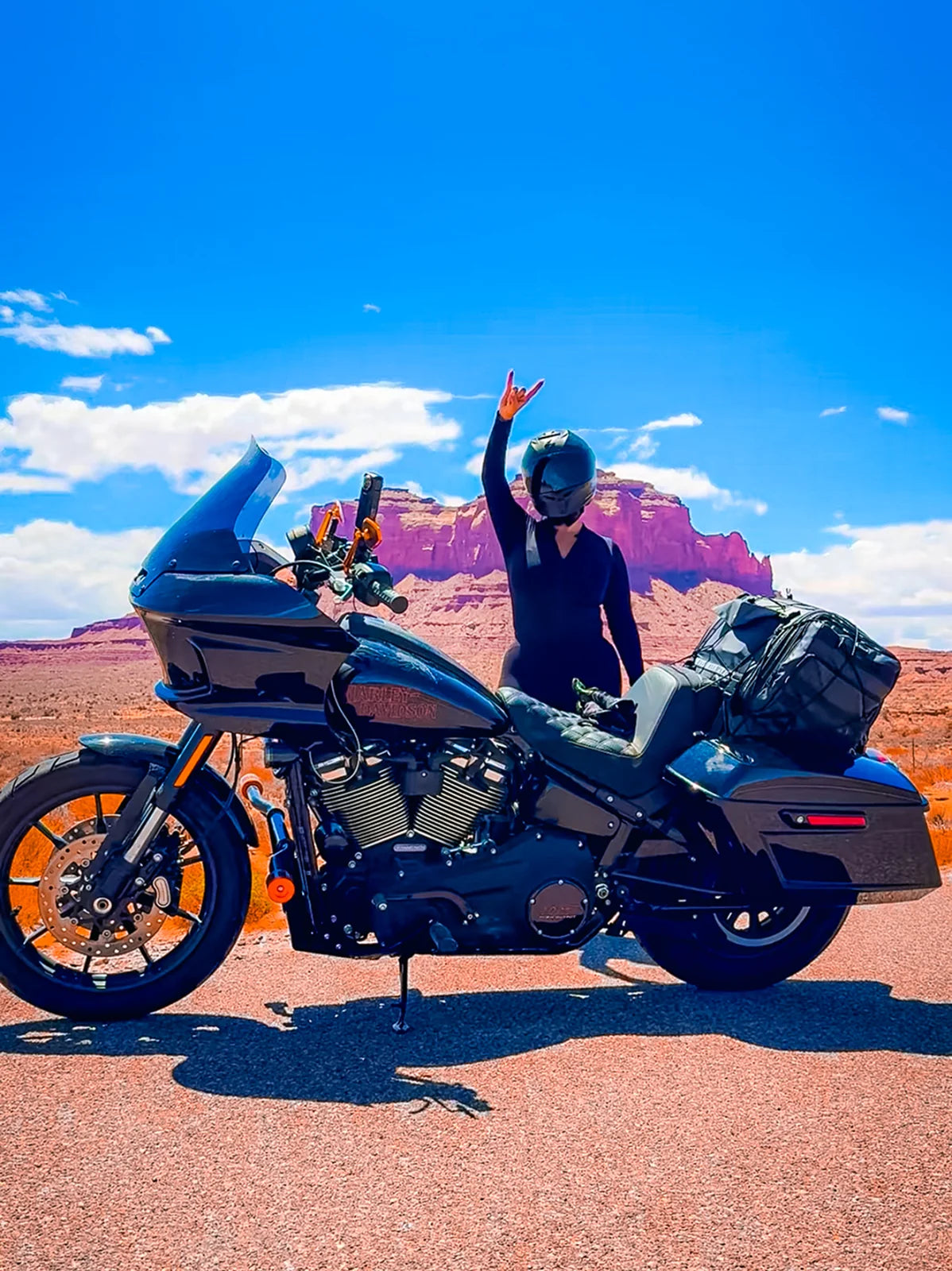 Mar Orts raising one hand beside a touring motorcycle in a red-rock desert landscape.